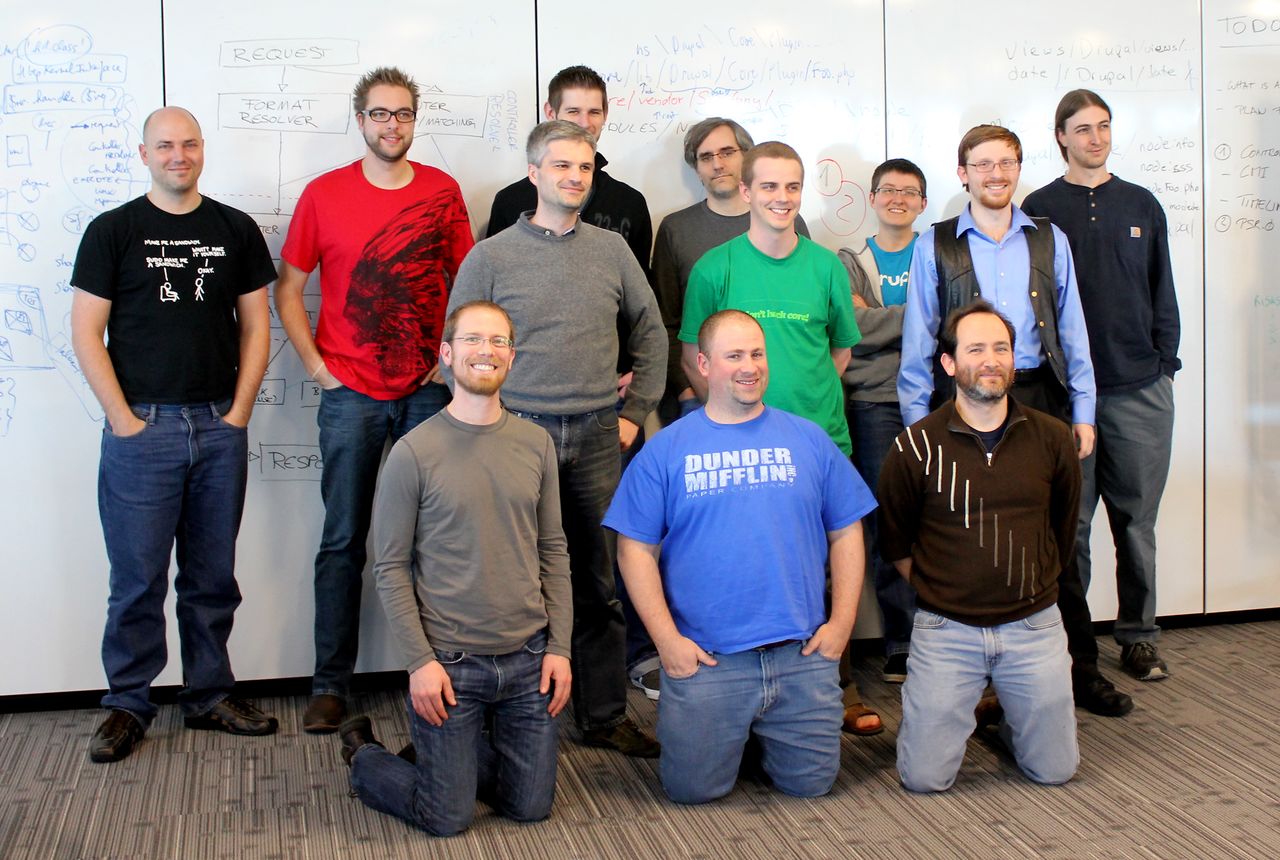 A group of eleven people posing for a photo in front of a whiteboard with notes and diagrams.