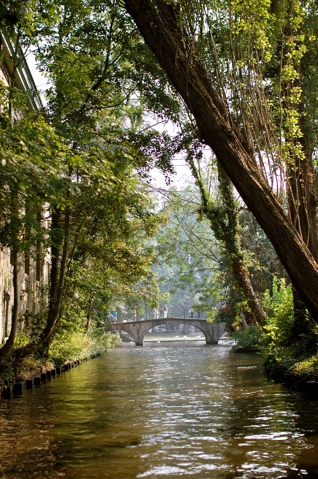 A canal in Bruges with a stone bridge, surrounded by green trees, and people walking across.