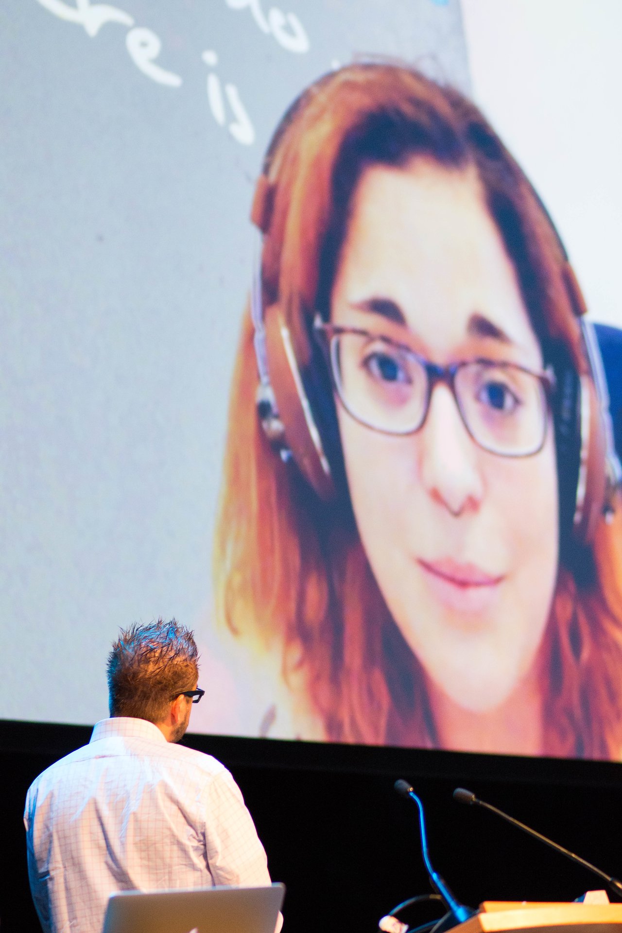 A speaker on stage looks at a large screen displaying a video of a woman presenting while wearing headphones.