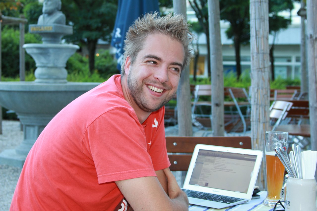 A smiling man in a red T-shirt sits at a beer garden table with a laptop and a glass of wheat beer.