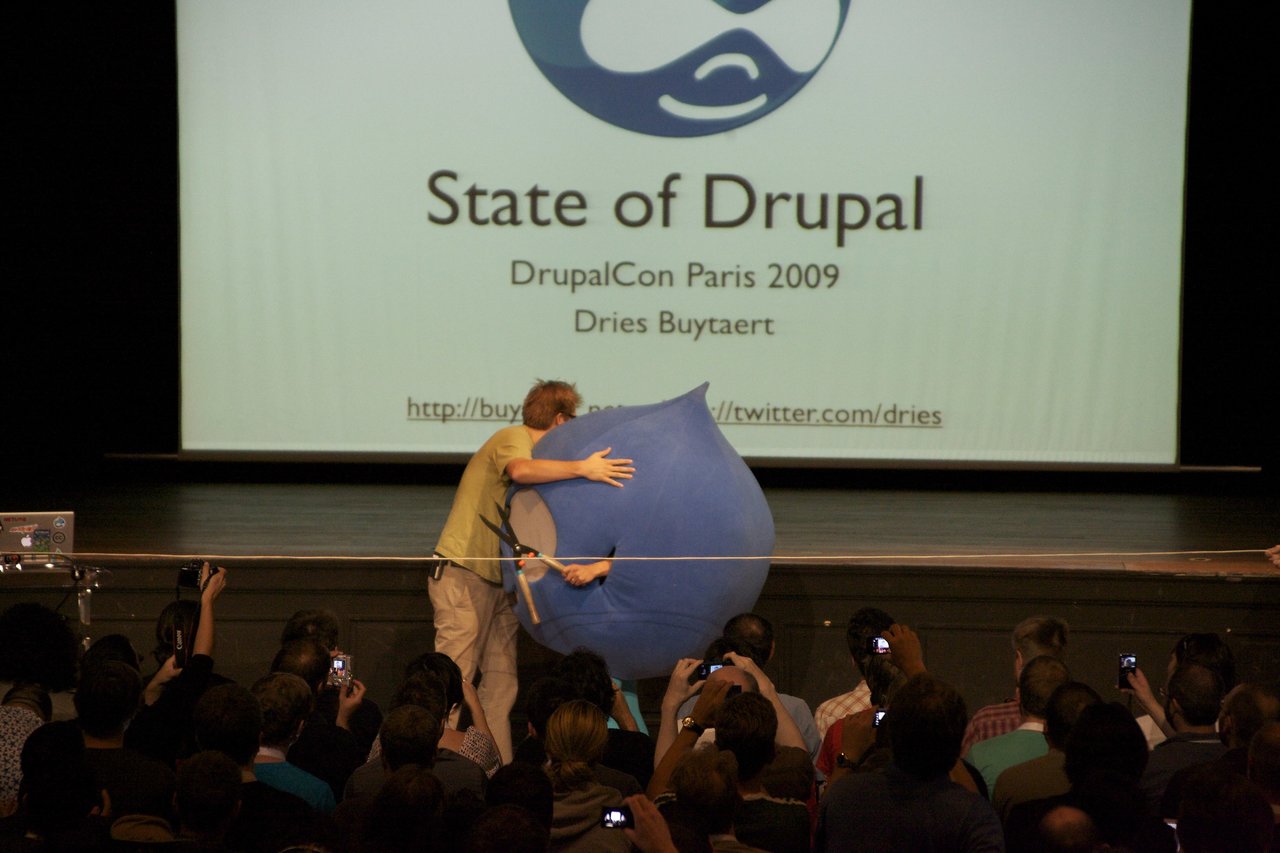 A speaker on stage hugs a large blue Drupal mascot while an audience takes photos at DrupalCon Paris 2009.