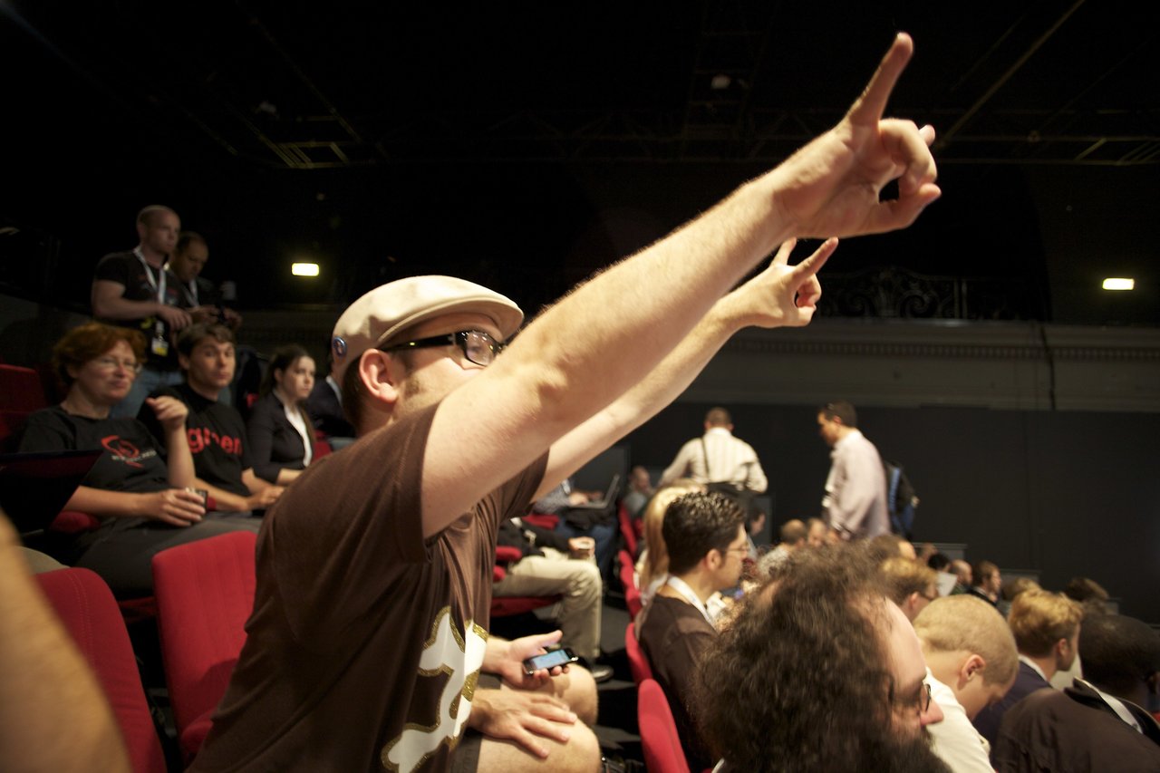 A man in a hat enthusiastically raises his hand in a crowded conference audience.