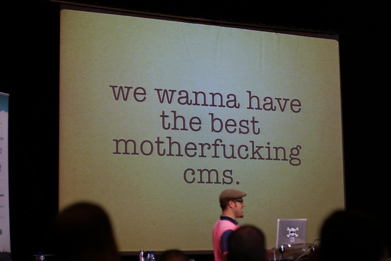 A speaker stands in front of a large screen displaying bold text about creating the best CMS at DrupalCon.