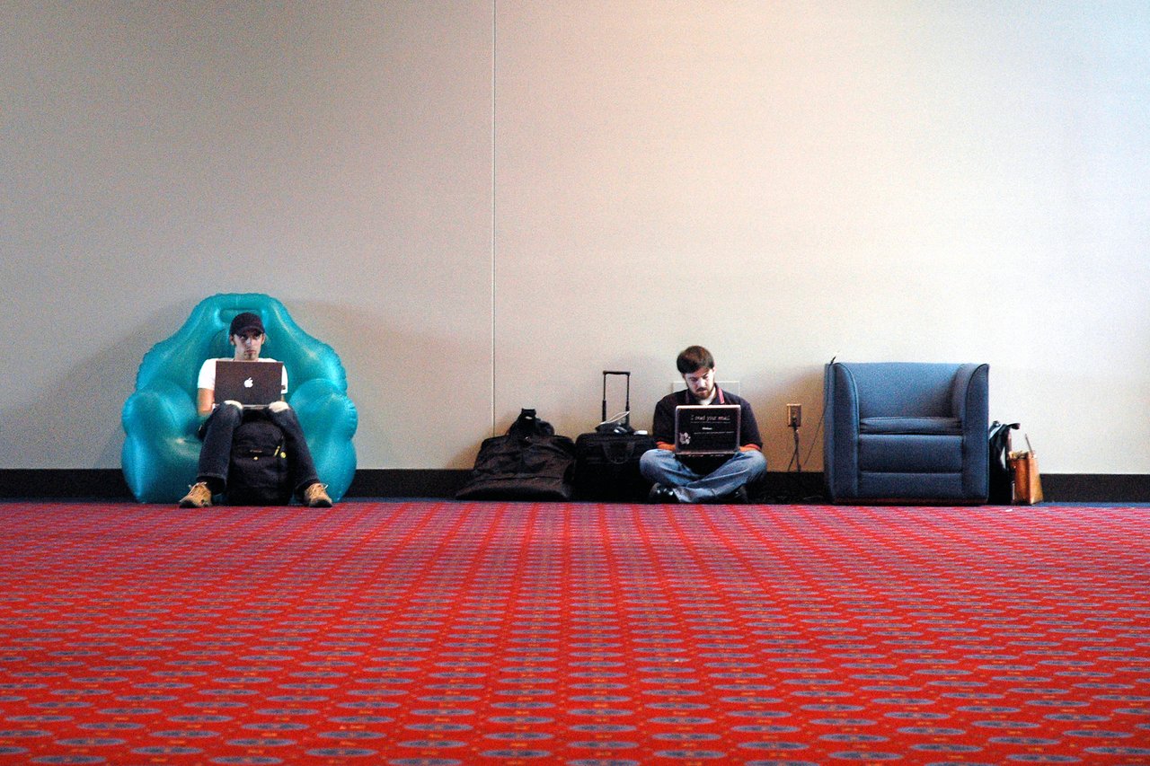 Two people sit against a wall in a convention center, working on laptops, surrounded by bags and chairs.