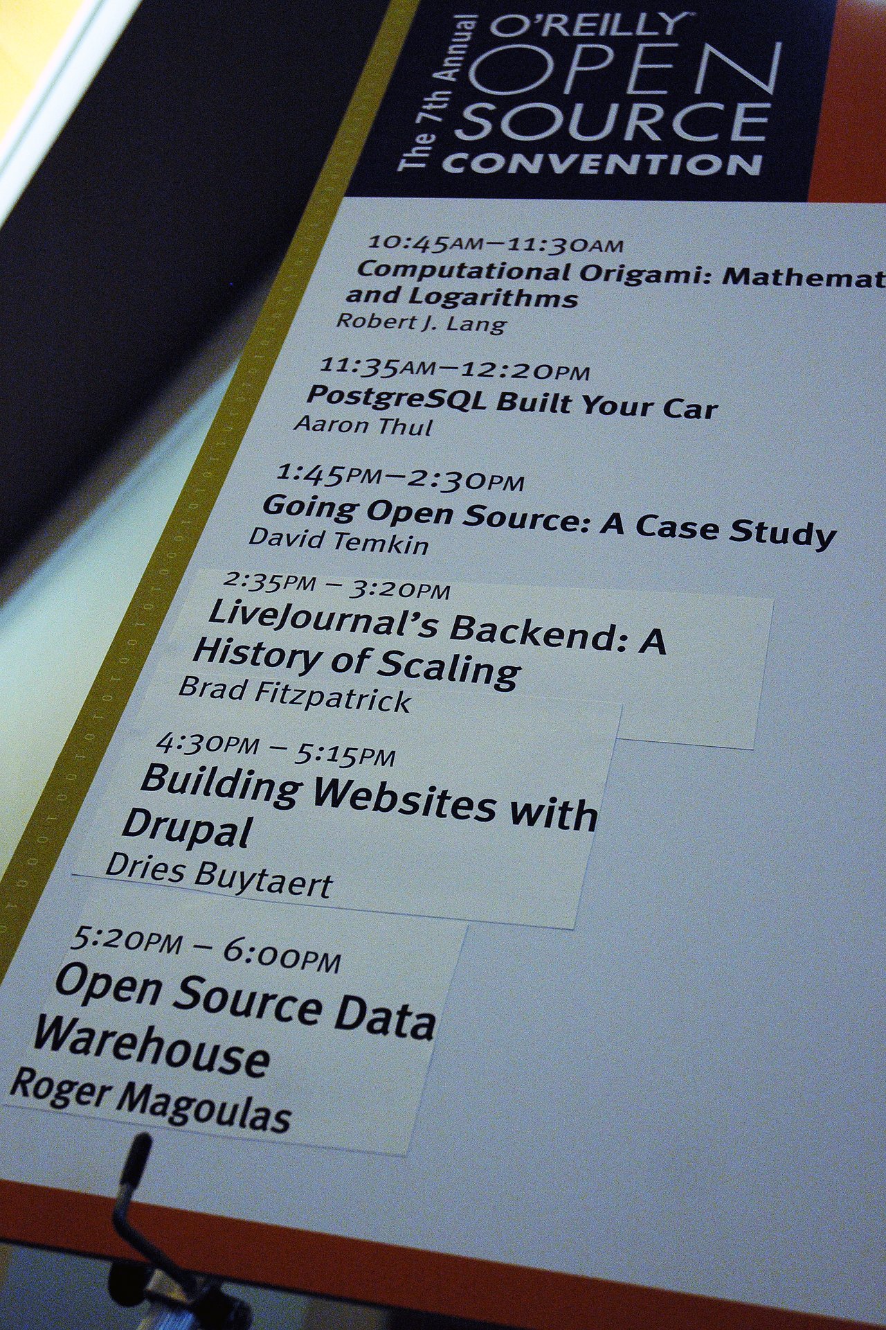 A conference schedule board displays session titles, including "Building Websites with Drupal" by Dries Buytaert at an open-source event.