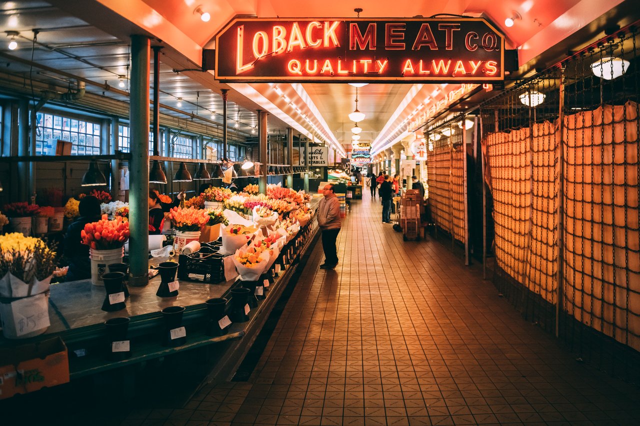 A person browses a flower stand inside Seattle's public market, with neon signs and vendors in the background.