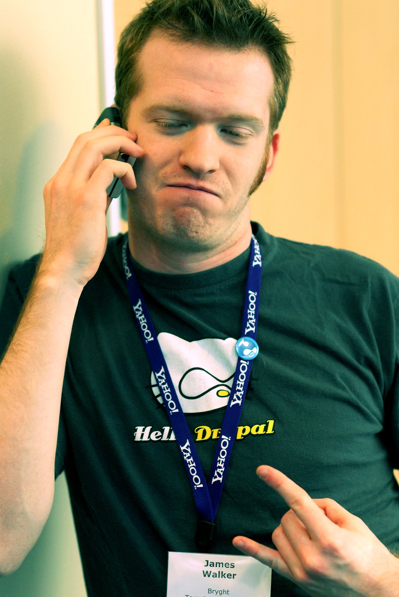 A man wearing a Drupal-themed shirt and Yahoo! lanyard talks on the phone while making a hand gesture.