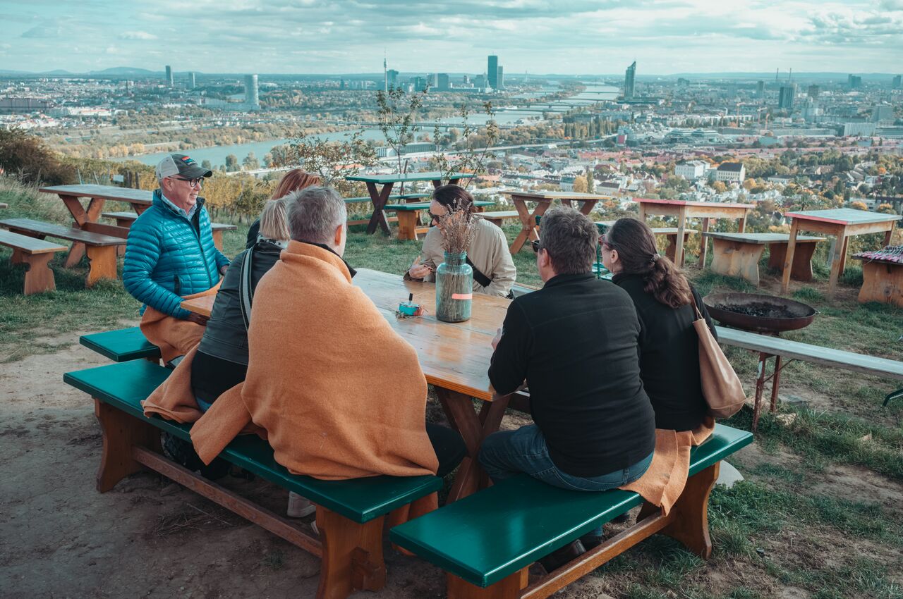 A group of friends sit around a picnic table in the hills above Vienna, wrapped in blankets and talking while enjoying the view.