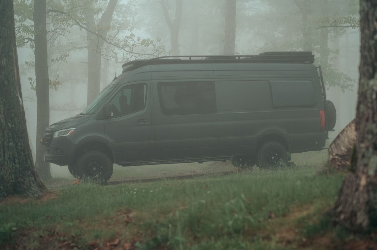 Matte black camper van parked among trees in dense morning fog.