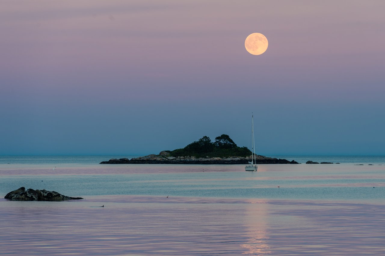 A full moon rises over calm ocean waters, with a small island and a sailboat in the distance.