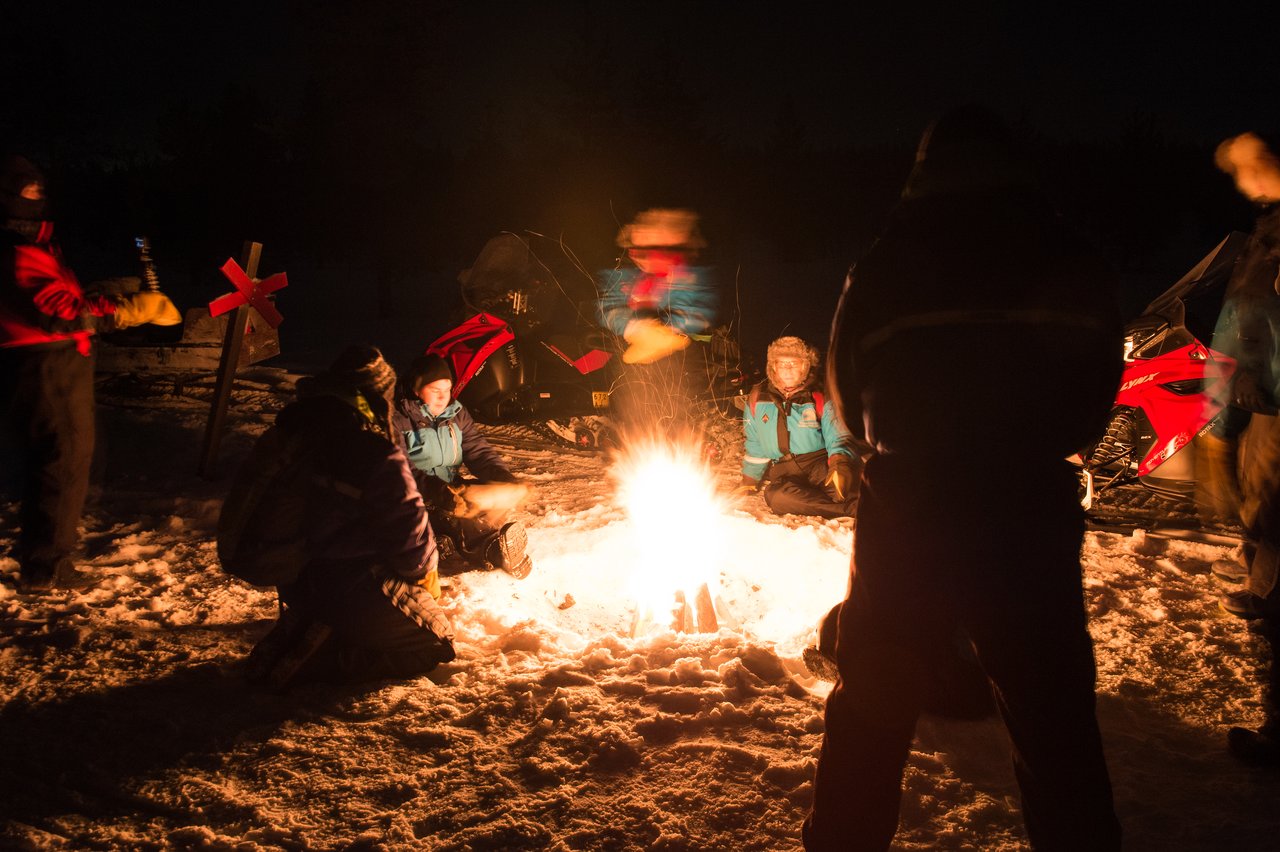 A group of people in winter clothing sit and stand around a campfire in the snow at night.