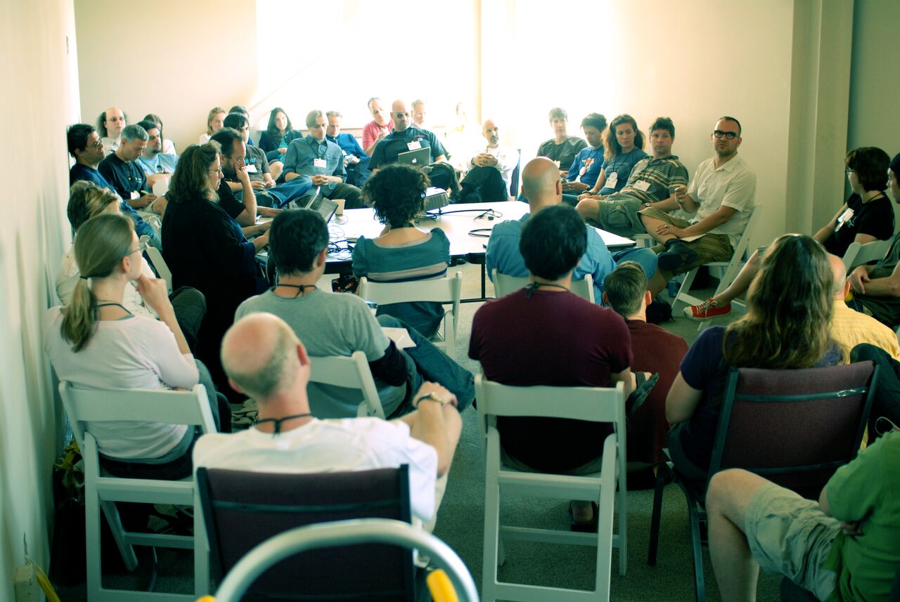 A group of people sit in a circle, attentively listening to a speaker in a discussion setting.