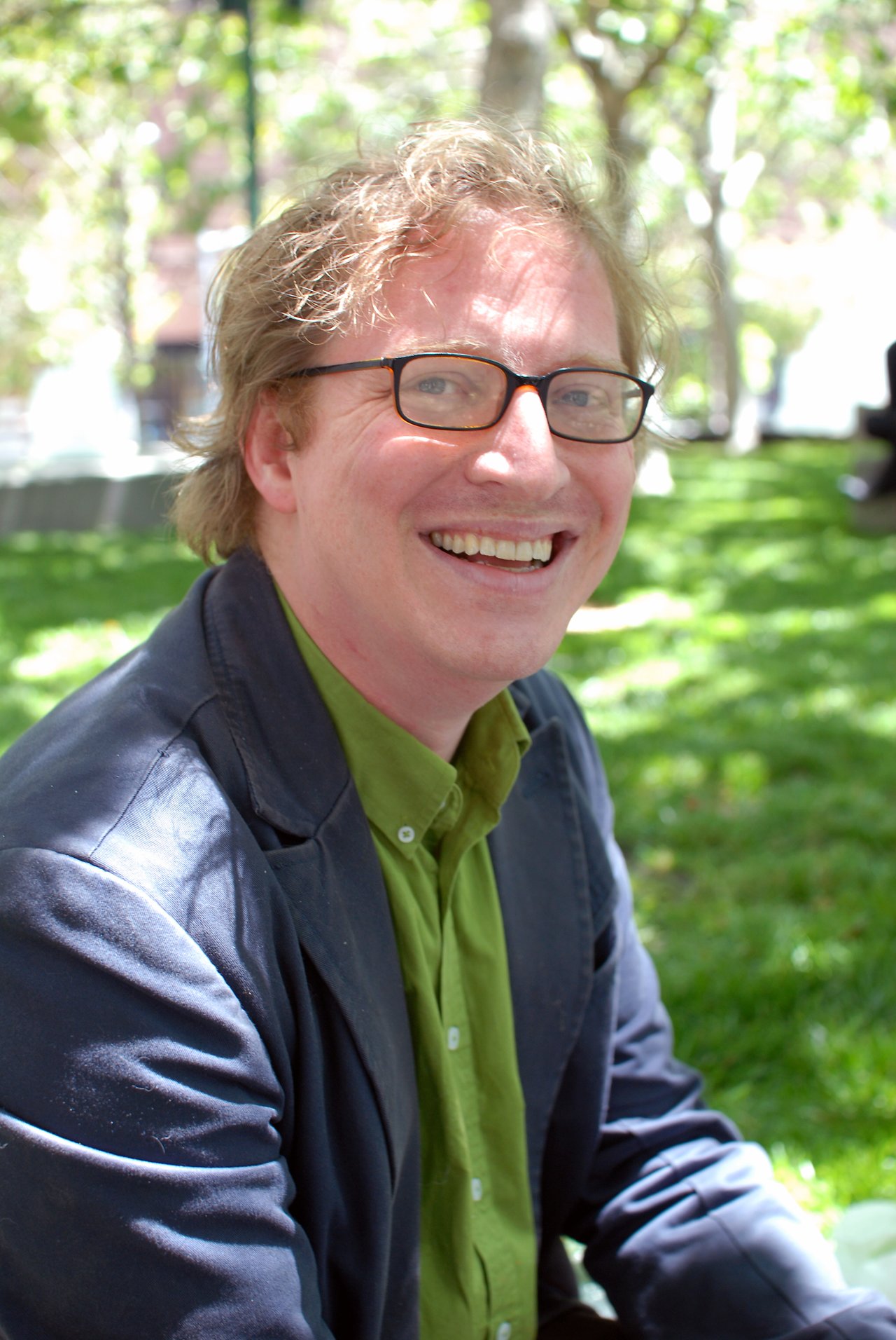 A man with glasses and a green shirt smiles while sitting outdoors on a sunny day.