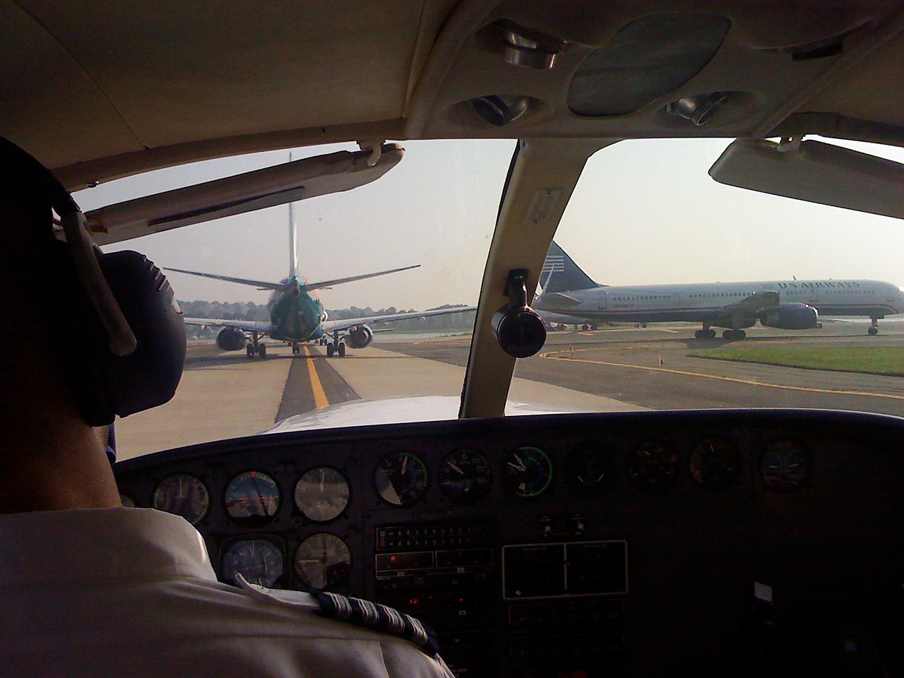 View from inside a small airplane cockpit, following a larger plane on the taxiway, with a pilot in the foreground.