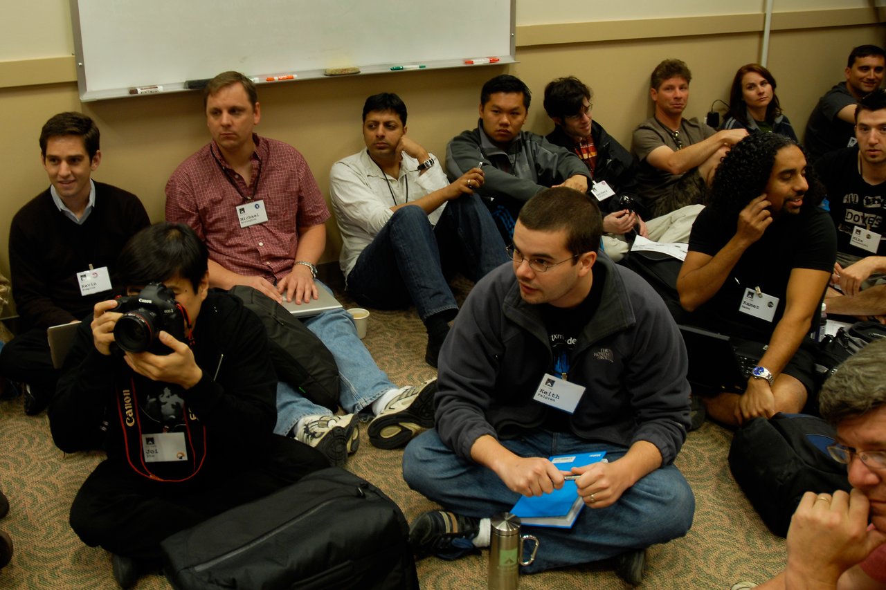 A person in a black hoodie is sitting on the floor, holding a camera and taking a photo in a group discussion.