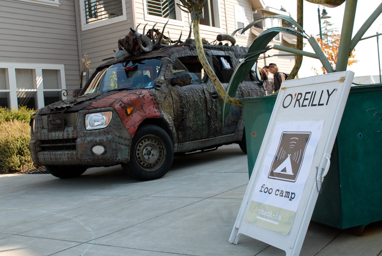 A heavily modified, rusted vehicle is parked near a "Foo Camp" check-in sign outside a building.