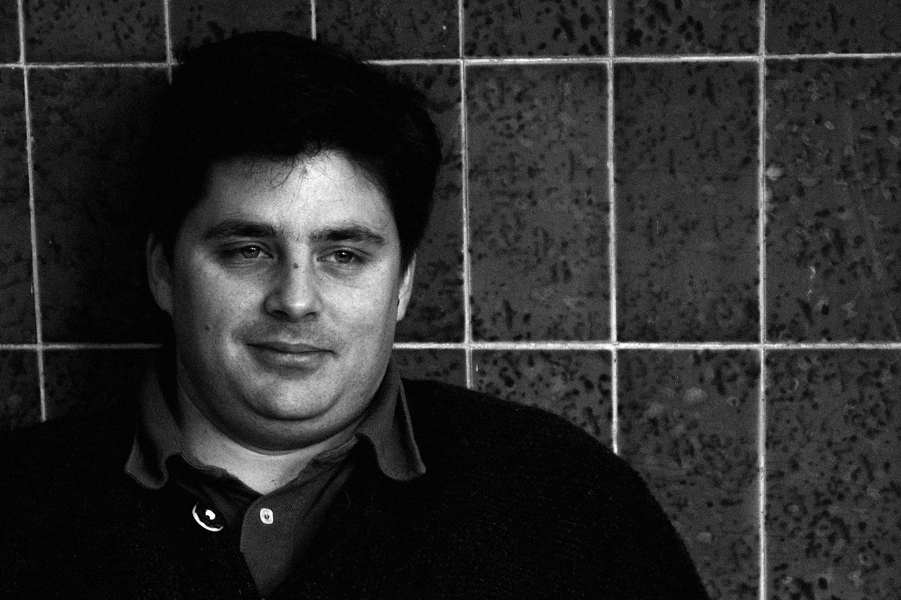 A man with short dark hair leans against a tiled wall, looking slightly to the side with a neutral expression.