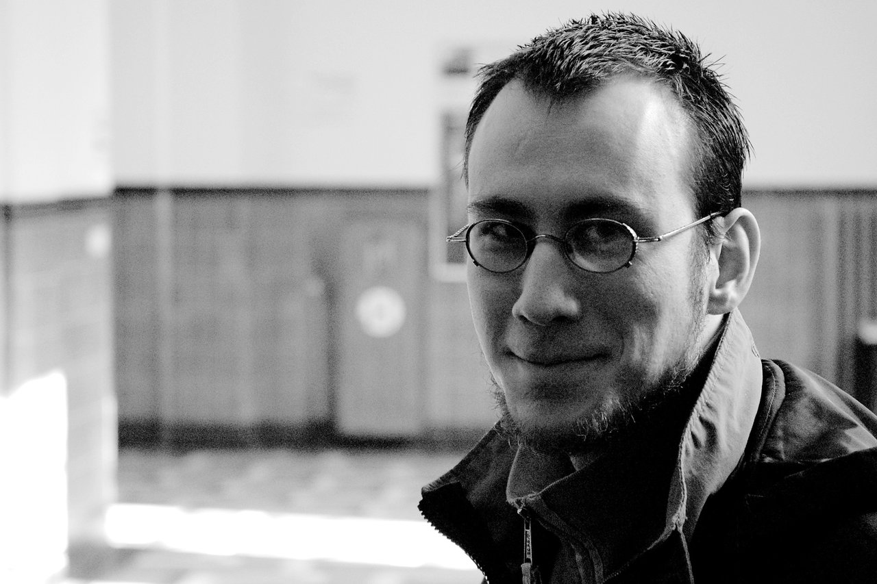 A man with glasses and a beard smiles slightly while looking at the camera in an indoor setting.