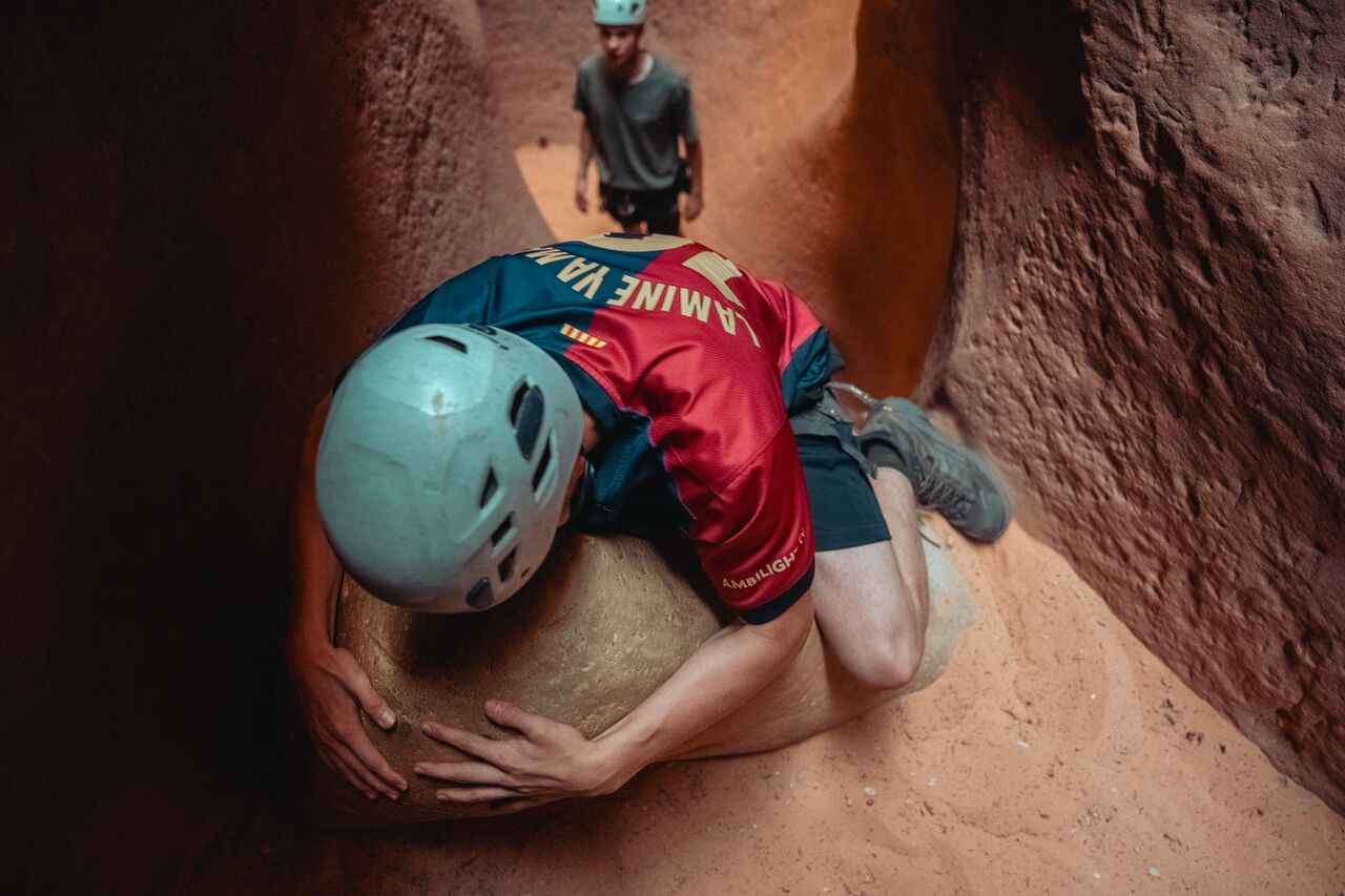 A teenager wearing a helmet lowering himself down while holding onto a large boulder inside a canyon in Zion National Park.