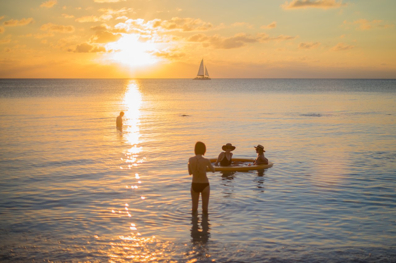 People wade in the ocean while others float on an inflatable, with the sun setting and a sailboat nearby.