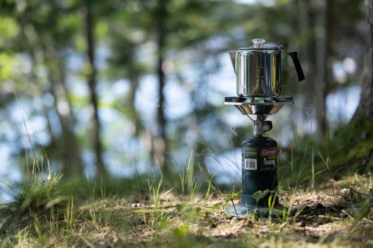 A moka pot brewing coffee on a portable camping stove in a forest setting.