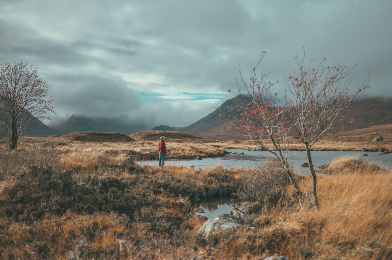 A person standing by a lake, surrounded by golden grass and mountains in the background.