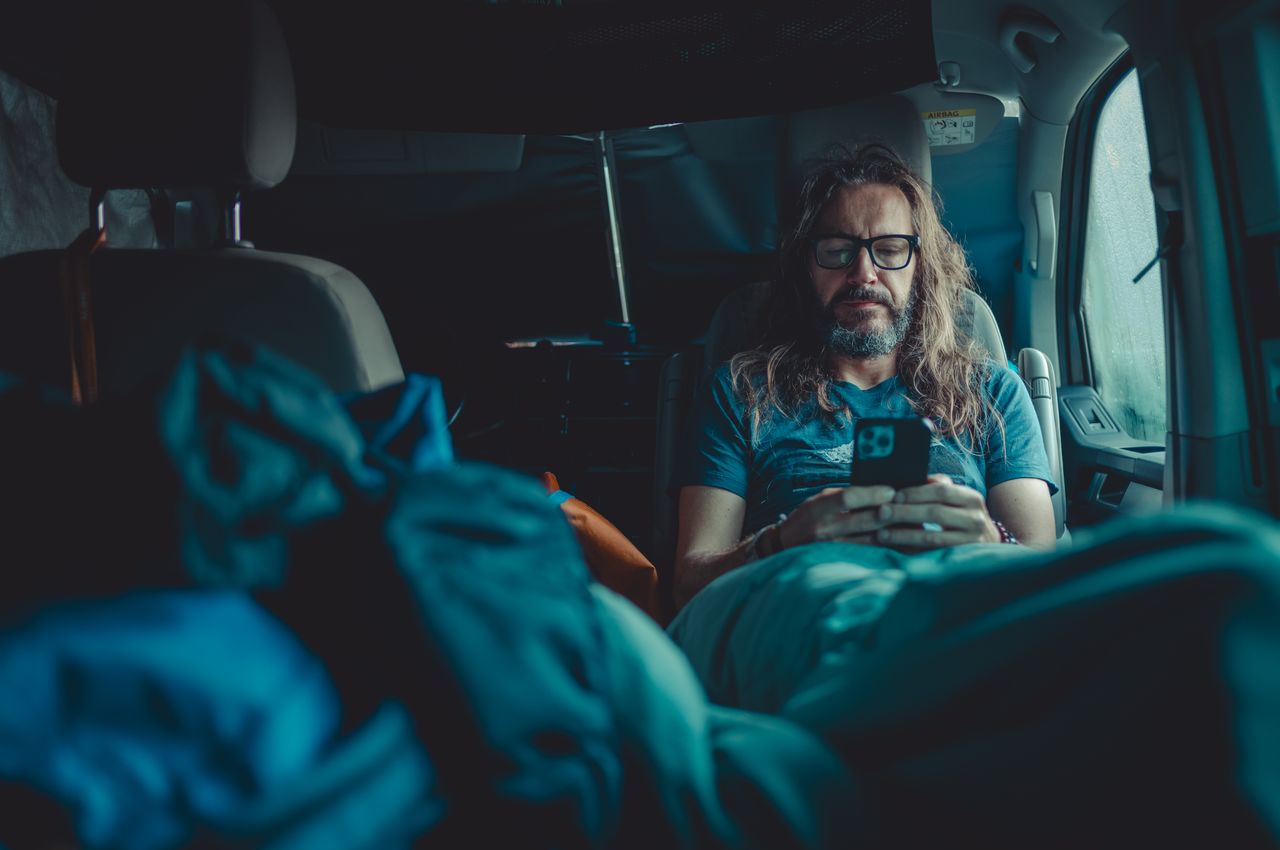 A person with long hair, sitting in a van, using a smartphone after just waking up.