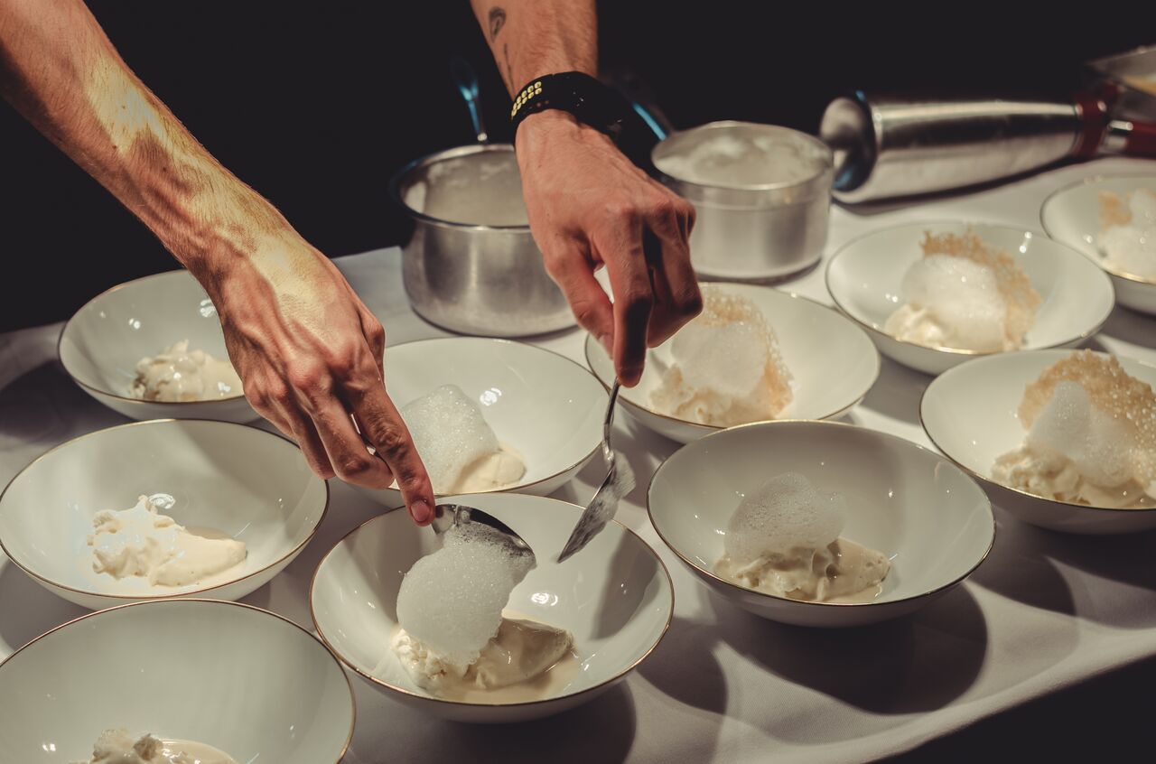 A chef's hands plate rows of white bowls of Parmigiano cream, each topped with an airy foam and crisps, on a white tablecloth.