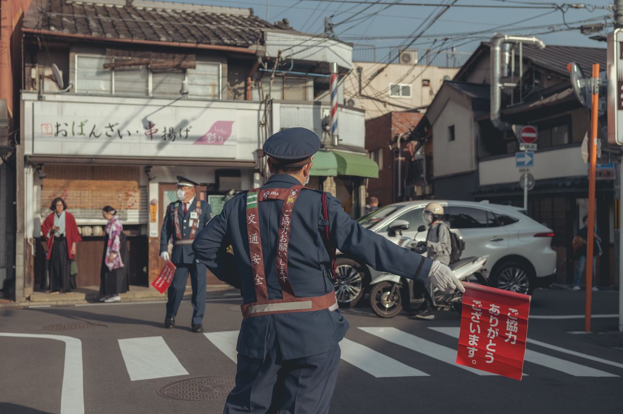 A traffic guard in a dark blue uniform, holding a red flag with white Japanese script, directs traffic on a busy Kyoto street.