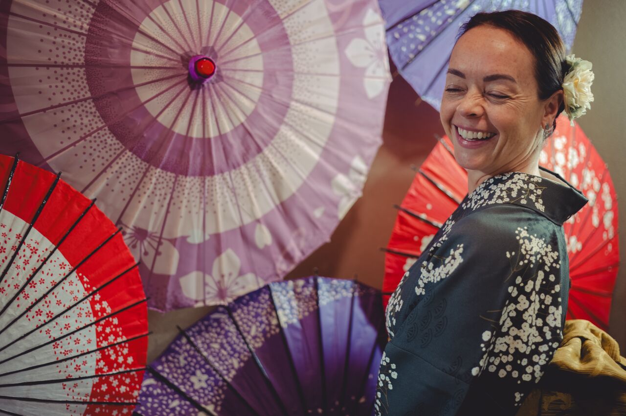 A woman wearing a traditional kimono smiles while standing in front of colorful paper umbrellas during a tea ceremony in Tokyo, Japan.