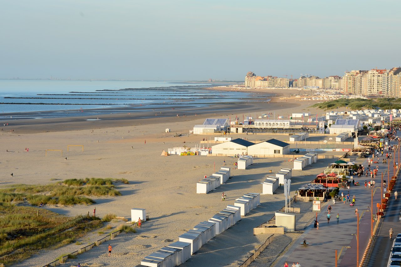 A sandy beach with small white beach huts, people walking, and a boardwalk next to the shoreline.