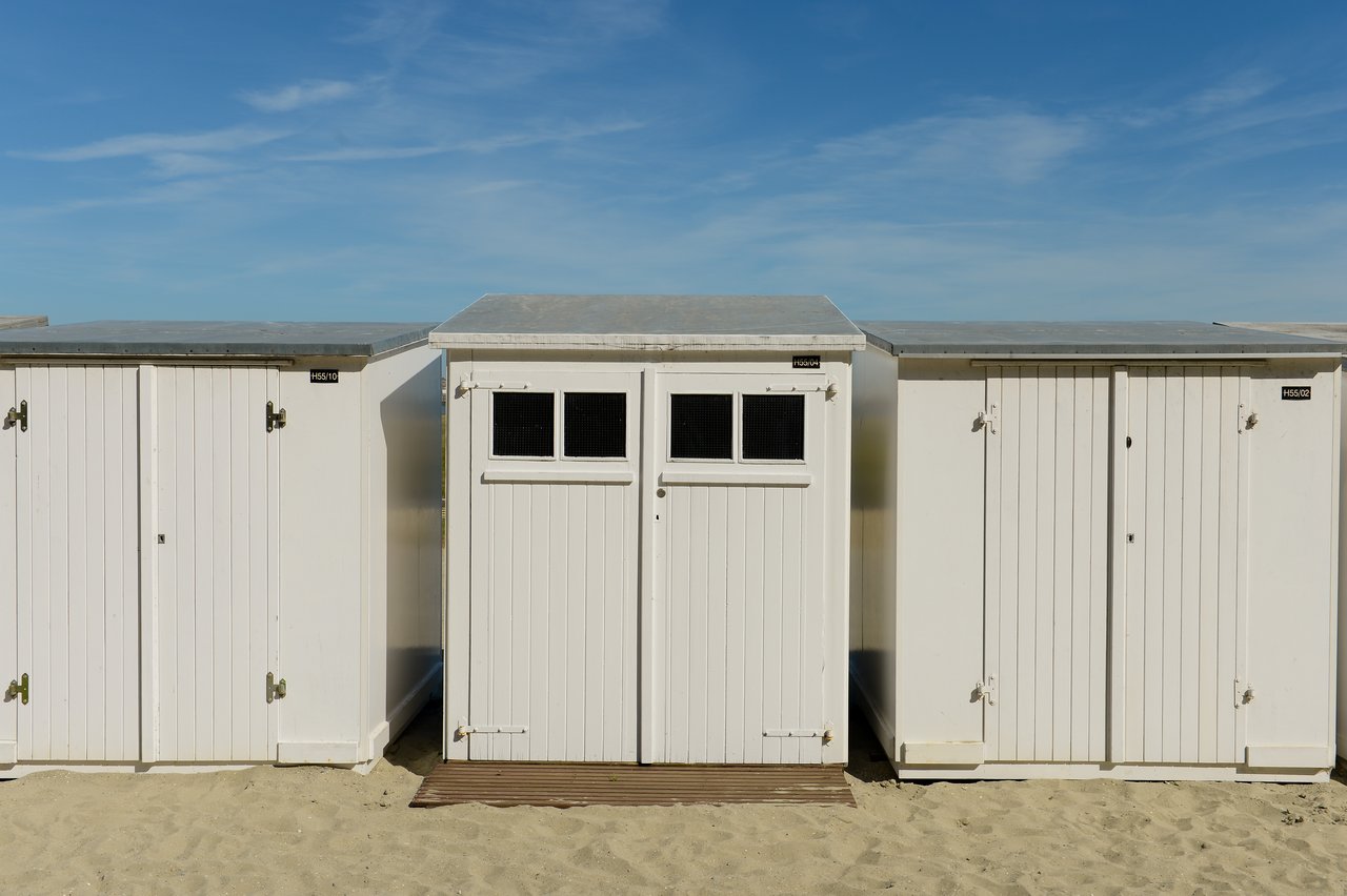 A row of white beach cabins with closed doors, standing on sandy ground under a clear blue sky.