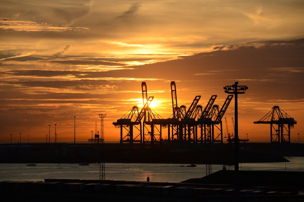 Silhouetted cranes stand against an orange sunset sky at a shipping port near the water.