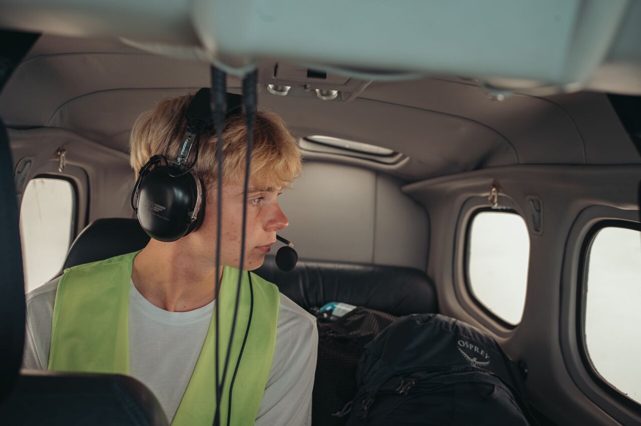 Passenger in a small plane wears a headset and safety vest, looking out the window, with a backpack beside him.