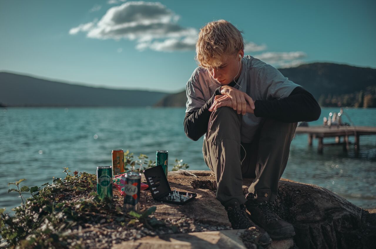 A young person sits on a lakeside rock, studying a small travel chess board and thinking about his next move.