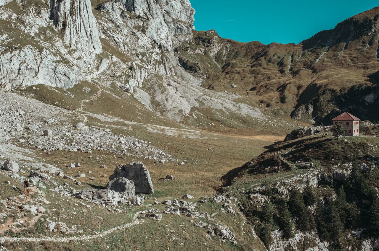 An old mountain hut stands on a hillside, and a narrow trail winds through the rocky meadows below where two hikers are descending.