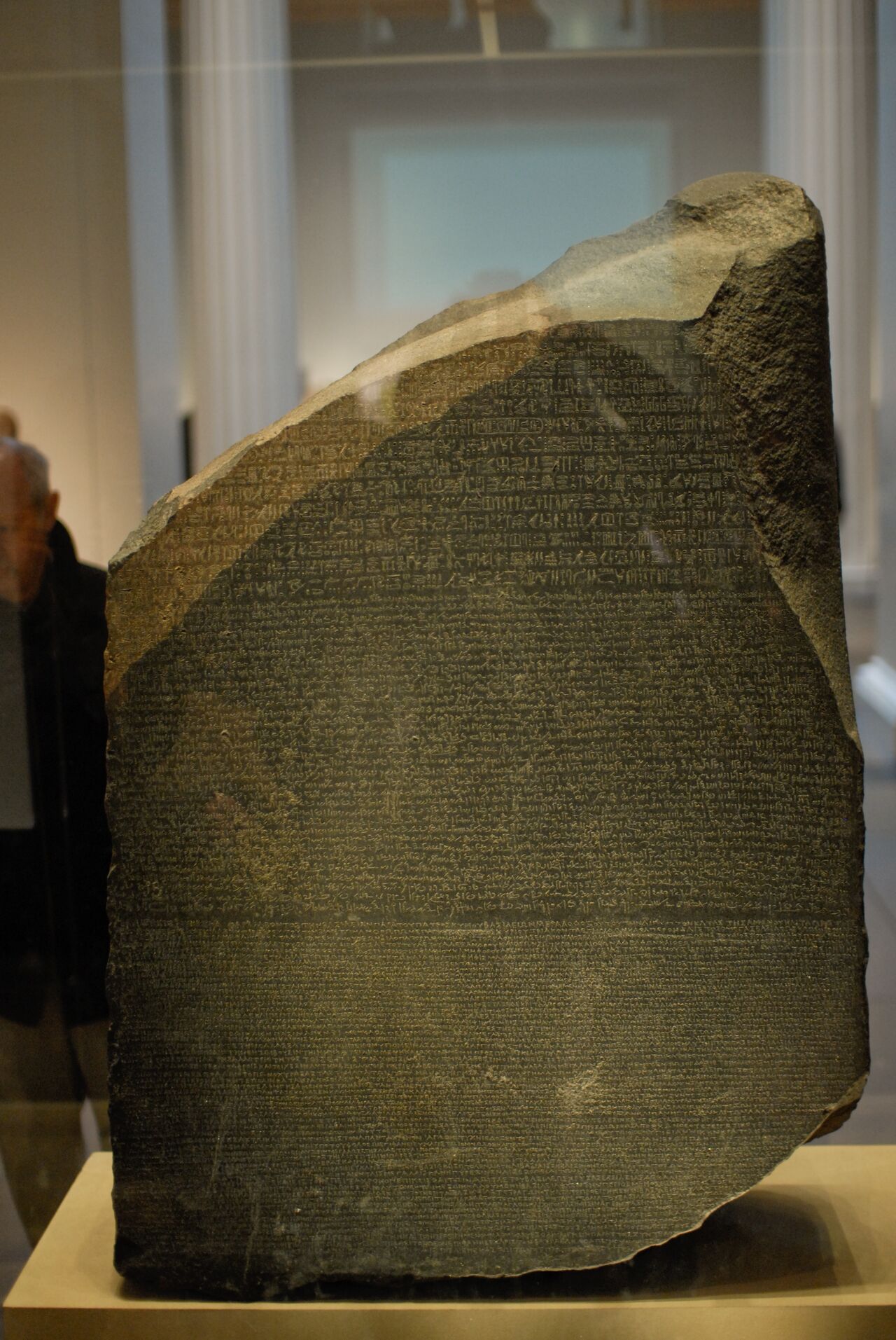 The Rosetta Stone, an ancient inscribed slab with three scripts, displayed in a museum with visitors in the background.