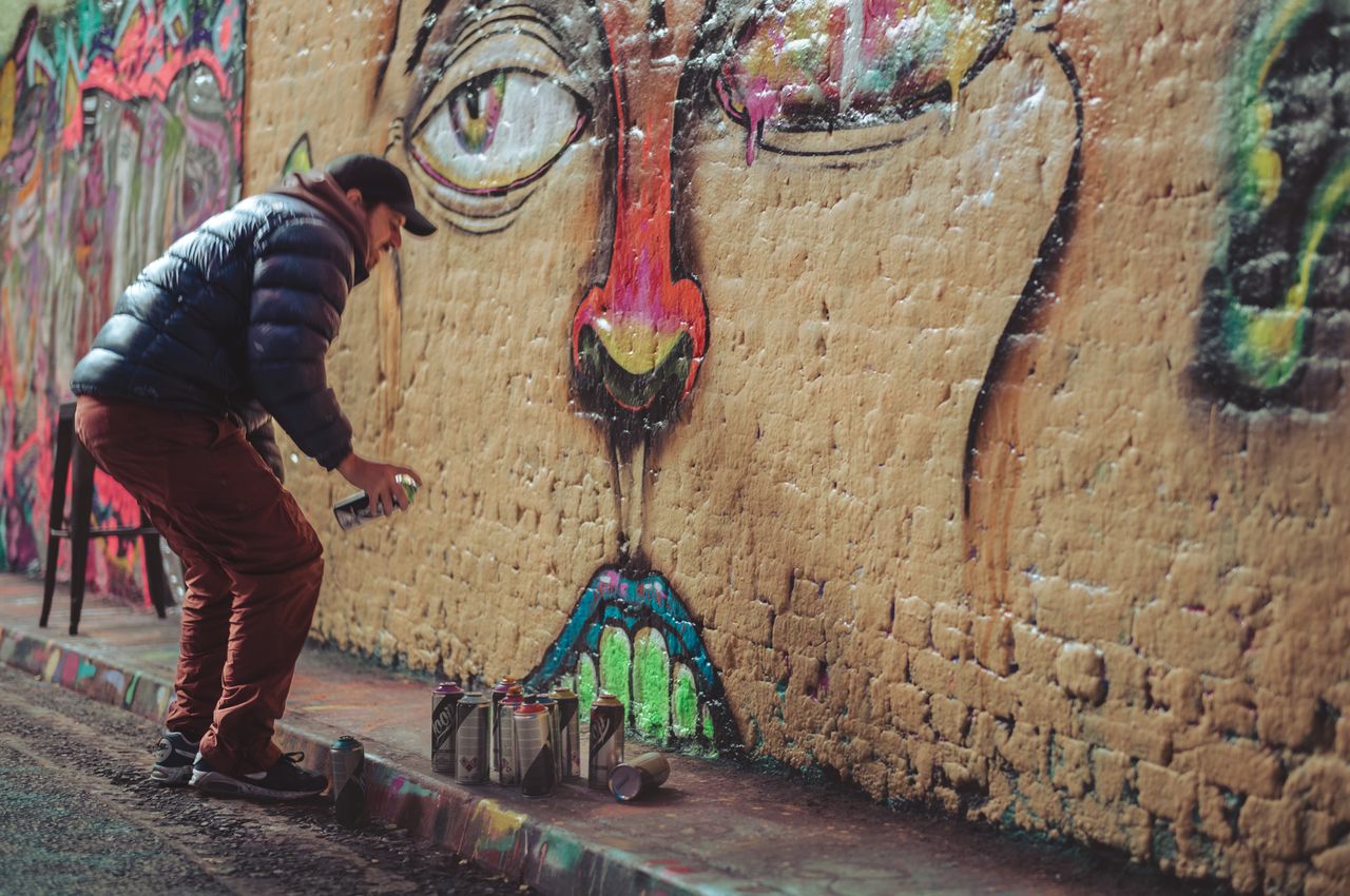 A man wearing a hat is spray-painting a wall with multiple cans of different colored paint at his feet.