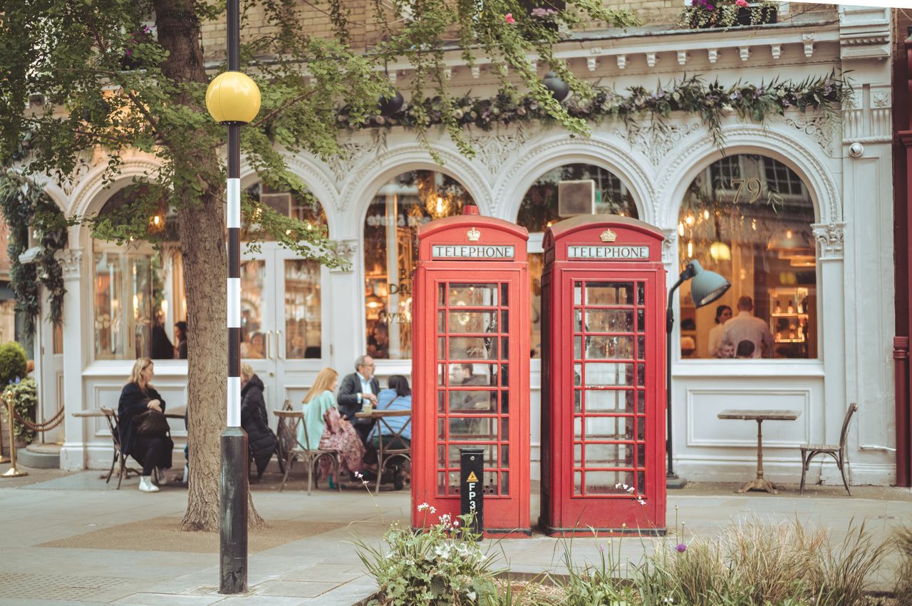 The foreground shows two red London phone booths while the background shows people sitting at outdoor tables in front of a restaurant.
