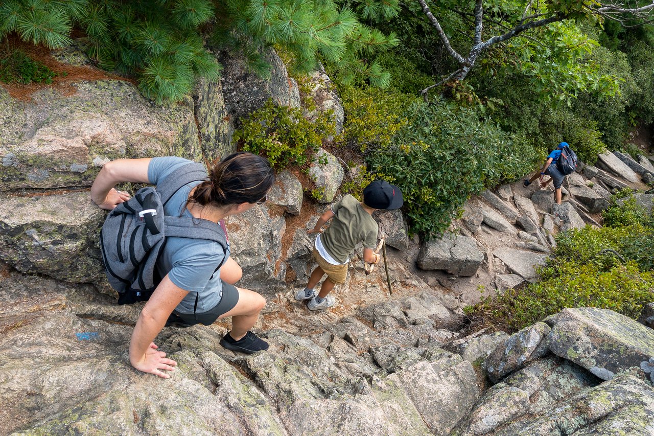 Three hikers carefully climb down a steep, rocky trail, using their hands and walking sticks for support.