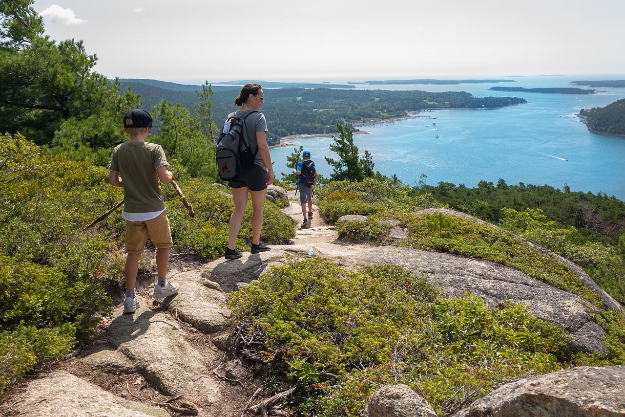 Three people hike down a rocky trail with a scenic view of water, islands, and greenery in the distance.