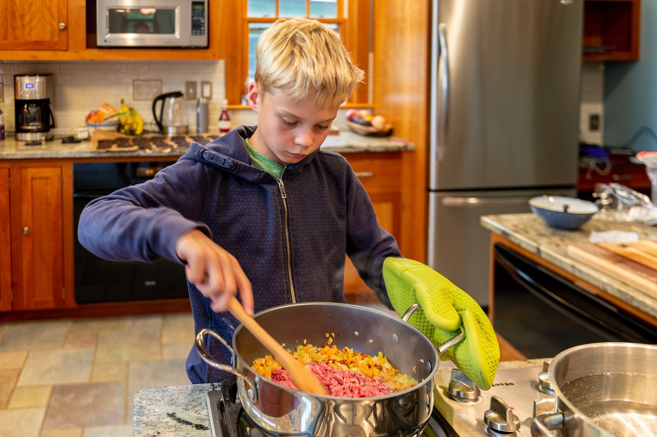 A young boy stirs ground meat and vegetables in a pot on the stove while wearing an oven mitt.