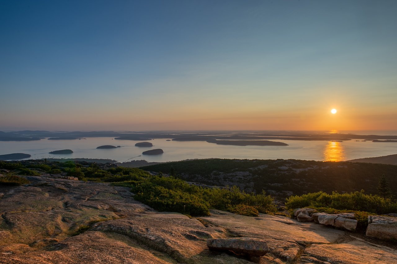 The sun rises over distant islands and calm water, viewed from a rocky hilltop with green vegetation.