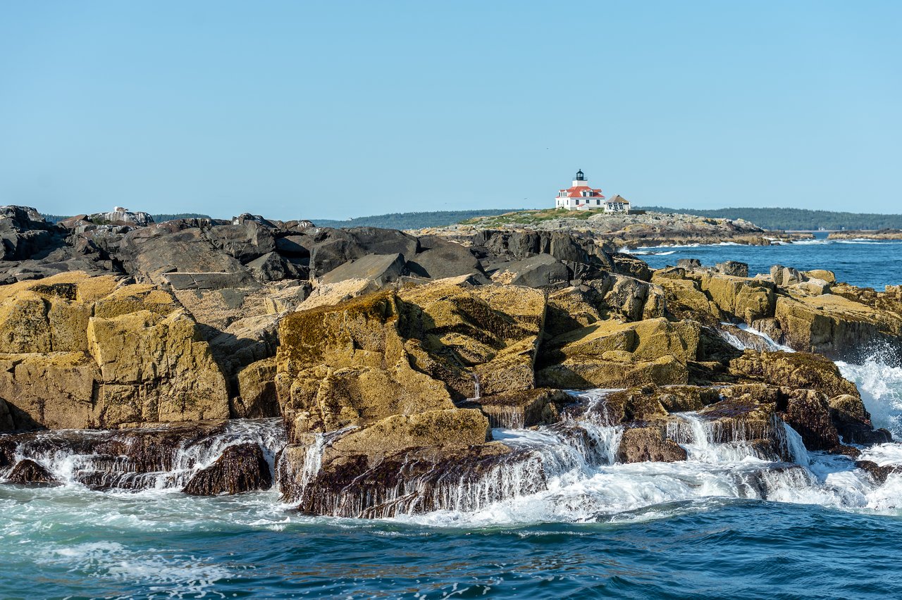 Waves crash against rocky shore with a white lighthouse in the distance under a clear blue sky.