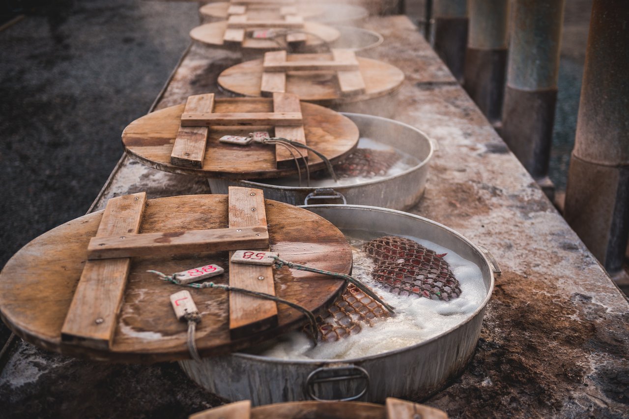 Several metal pots with wooden lids boil ingredients in hot water, with steam rising from the surface.