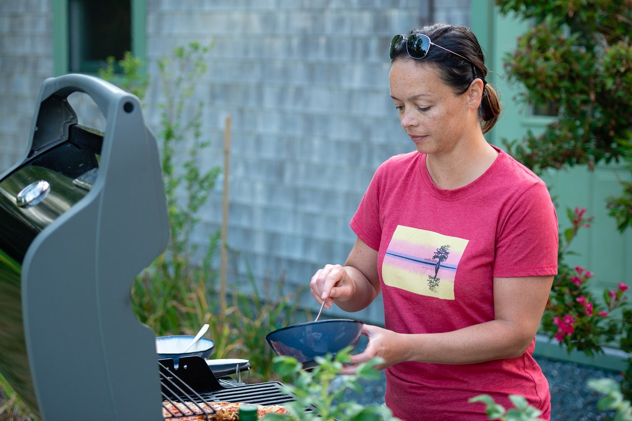 A woman in a red shirt prepares food at a grill, holding a bowl and stirring with a spoon.