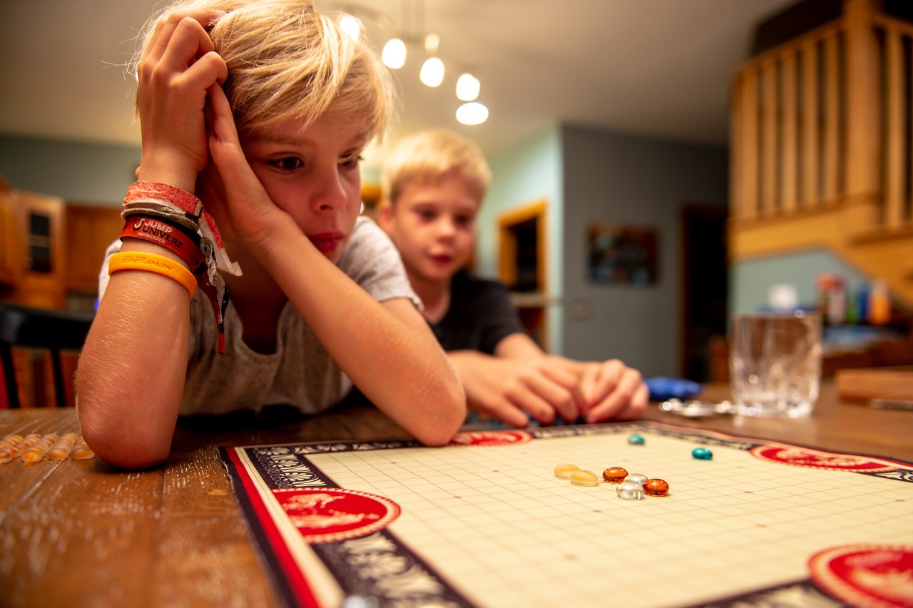 Two children sit at a table, focused on a board game with colorful glass pieces.