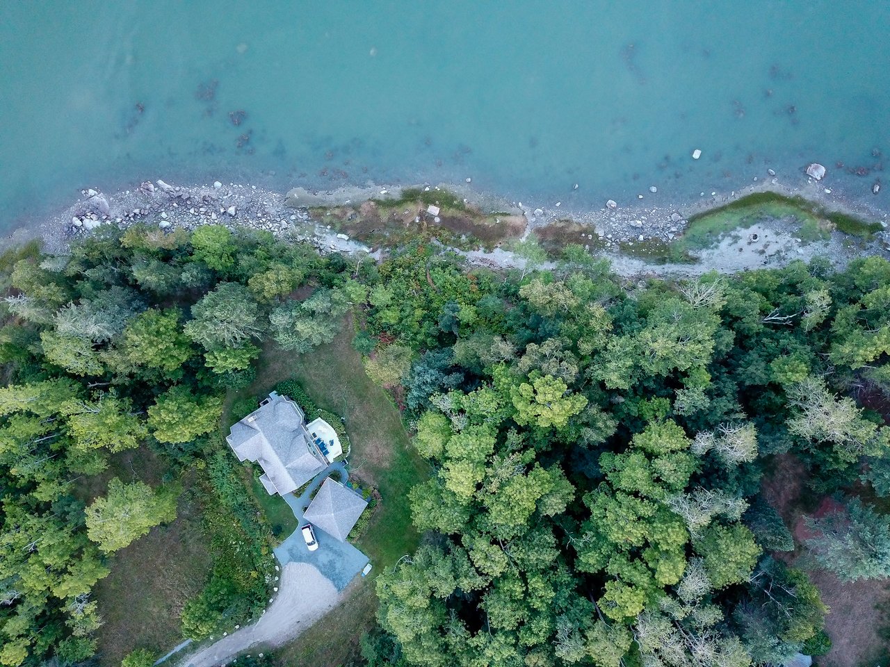 A house with a driveway and parked car is surrounded by trees near a rocky shoreline and calm water.