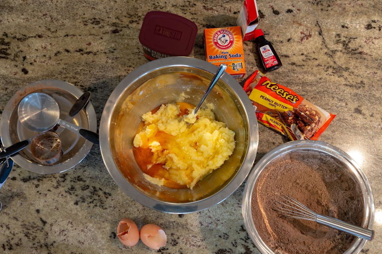 A bowl of creamed butter and sugar next to a bowl of cocoa and flour with a whisk, eggshells, baking soda and a bag of Reese's peanut butter chips on a granite counter.