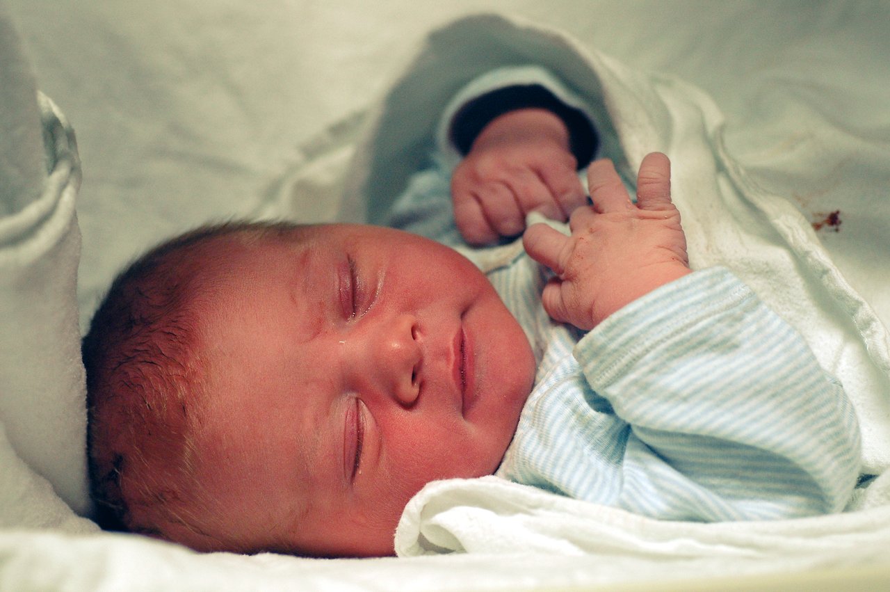 A newborn baby sleeps peacefully, wrapped in a soft blanket, with his hands resting near his face.